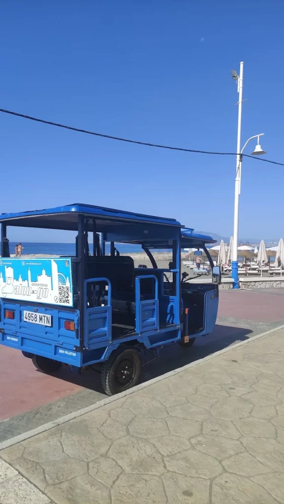 Tuk tuk azul de Baloo-Go rotulado con un código QR, aparcado junto a una playa con sombrillas