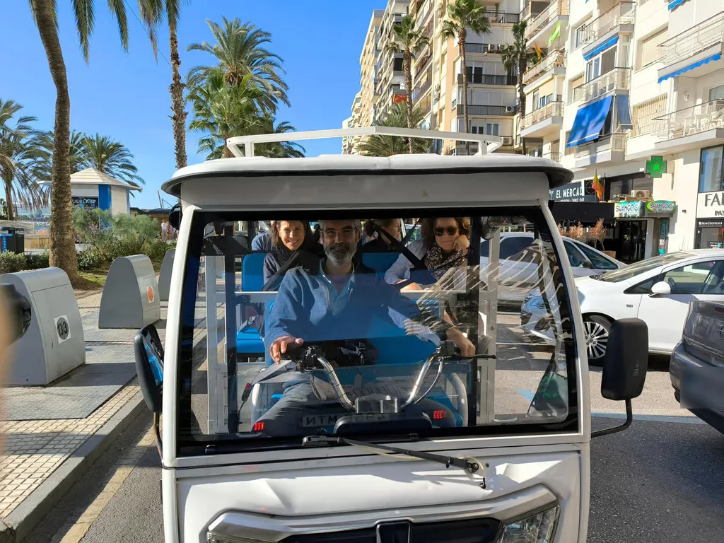 Tuk tuk blanco circulando por una calle con palmeras, con el conductor y varios pasajeros a bordo