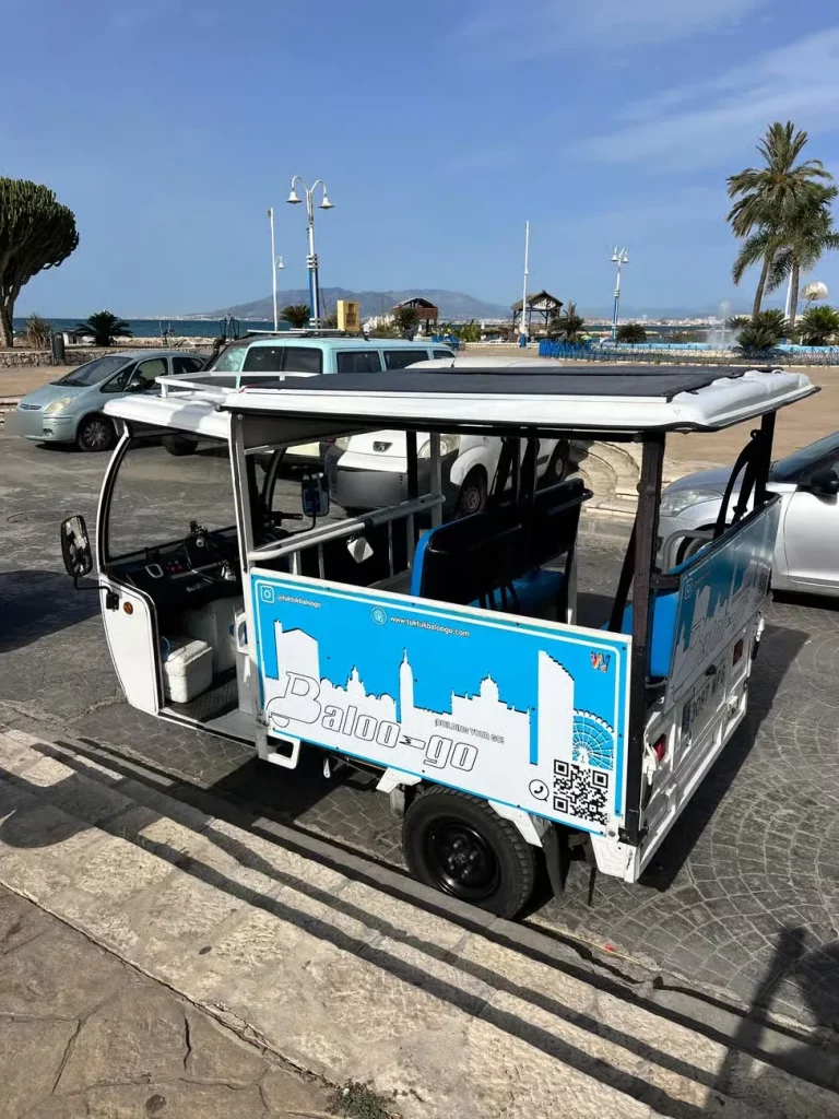 Tuk tuk blanco de Baloo-Go visto de lado, con el panel trasero rotulado con la silueta de una ciudad y un código QR