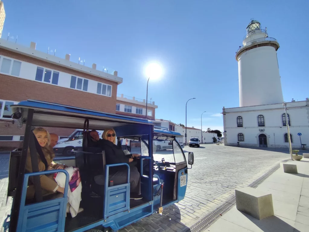 Tuk tuk azul de Baloo-Go con pasajeros sonrientes junto a La Farola de Málaga