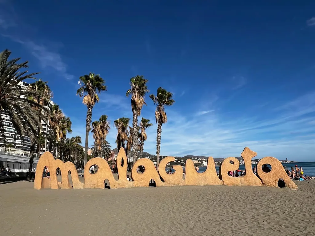 Escultura con las letras de 'Malagueta' en la arena de la playa de Málaga, junto a un grupo de palmeras